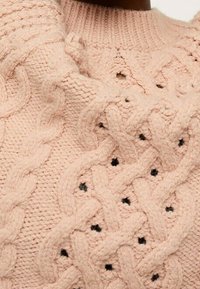 Gros plan sur un pull tricoté en gros mailles rose pâle avec des motifs en câble tressé et de petits accents de points noirs.