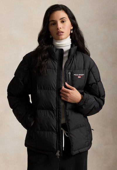 Juoda pūsta striukė su siūtu raštu, užtraukiamu priekiu ir aukštu apykakliu. Kairėje krūtinės pusėje yra „POLO SPORT“ logotipas, o rankogaliai yra elastingi.