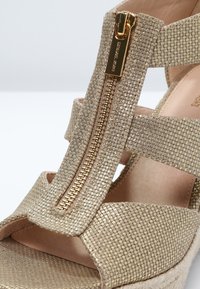 Sandale beige texturée avec une tirette de fermeture éclair dorée à l’avant gravée du nom de la marque et plusieurs larges brides sur le dessus du pied.