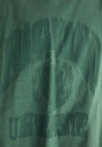 Camiseta de color verde oscuro con un gran estampado desvaído de "HARVARD" arqueándose sobre un emblema de escudo y "UBER" en texto negrita debajo.