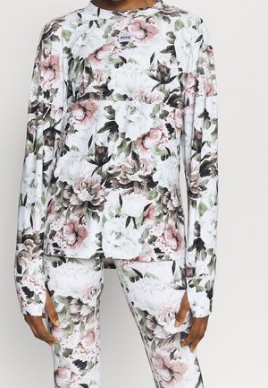 Osoba ubranа w dopasowany, długi rękaw i spodnie z nadrukiem kwiatowym o dużych białych, różowych i zielonych kwiatach na białym tle.
