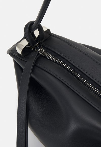 Sac à main en cuir noir avec fermeture éclair argentée et bandoulière attachée à une boucle métallique sur un fond clair uni.