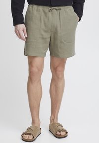 Shorts en coton texturé vert olive avec une taille élastique, un cordon de serrage et deux poches avant. Portés avec des sandales à glisser marron.