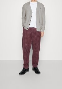 Ljusgrå cardigan med bruna knappar, vit t-shirt och bordeauxmönstrade byxor, matchat med svarta skor, stående mot en neutral bakgrund.