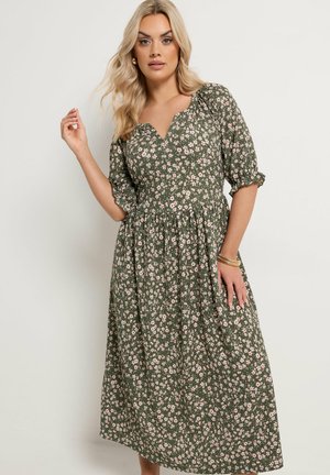 Kvinde iført en grøn midi-kjole med blomsterprint, puffede ærmer, V-udskæring og guldarmbånd på håndleddet, poserer mod en ensfarvet baggrund.