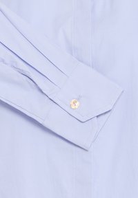Maniche di camicia azzurro chiaro con una texture liscia simile al cotone, con polsino a punta e un accento di bottone dorato.