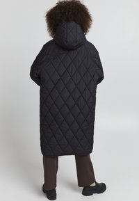 Manteau matelassé noir avec capuche, arborant un motif en losange. Le manteau a une coupe ample, atteignant le milieu du mollet, associé à un pantalon sombre et des bottes.