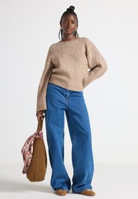 Pullover beige lavorato a maglia con motivo a trecce, maniche ampie; abbinato a jeans a gamba larga blu a vita alta. Borsa in suede marrone con un accento di sciarpa floreale.