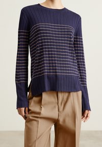 Granatowy sweter z długim rękawem, żebrowany, z poziomymi beżowymi paskami, okrągły dekolt i bocznymi rozcięciami; połączony z jasnobrązowymi spodniami.