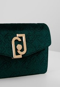 Pochette en velours texturé vert foncé avec un motif géométrique en relief et un fermoir décoratif doré sur un fond neutre.