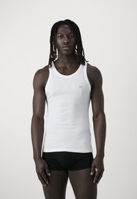 White cotton tank top with a round neckline, featuring contrast stitching and a small brand label on the left chest. Model wearing black shorts.