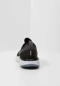 Zapato deportivo negro con parte superior texturizada, suela acolchada y detalles reflectantes. Presenta un diseño minimalista y un talón acolchado para mayor soporte.