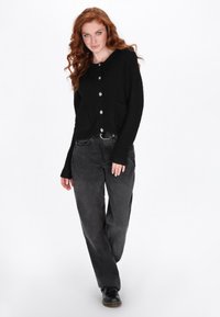Cardigan negru cu nasturi, dintr-un material texturat, având două buzunare în față și un tiv ușor desfăcut, asortat cu pantaloni gri închis.