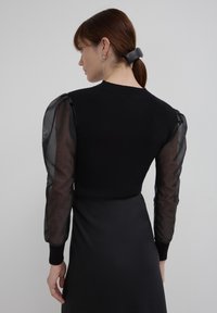 Donna con capelli castani raccolti con una clip nera, indossa un abito nero con maniche a sbuffo trasparenti, mostrata di spalle su uno sfondo grigio semplice.
