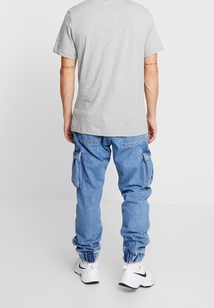 Mężczyzna w jasnoszarej koszulce, niebieskich dżinsach typu cargo z elastycznymi mankietami, białych tenisówkach, stojący tyłem na jednolitym białym tle.