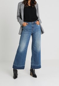 Jeans in denim blu a gamba larga con un lavaggio sbiadito e cuciture a contrasto, abbinati a un top nero e un blazer a quadri grigi. Stivali neri alla caviglia completano il look.