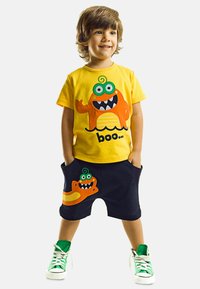 Gul bomulds t-shirt med et tegneserie monster grafik. Mørke shorts med en orange monster applikation. Grønne sneakers med hvide såler.