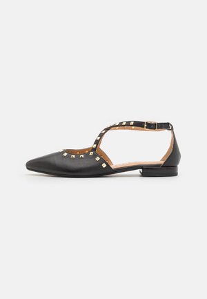 Zapato plano negro con punta afilada, correa de tobillo y acentos de tachuelas doradas en el empeine. Hecho de cuero suave con un tacón bajo.