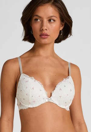 Vrouw draagt een witte kanten beha met kleine roze bloemenpatronen en zilveren verstelbare bandjes, kijkt iets opzij tegen een effen achtergrond.