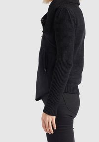 Personne portant un pull noir texturé en maille avec un gilet noir zippé par-dessus, vue de profil sur un fond clair uni.
