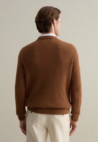 Brown knit sweater with a textured pattern, ribbed cuffs, and hem. Worn over light-colored trousers, viewed from the back.