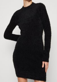 Kobieta w dopasowanej, czarnej, miękkiej, dzianinowej sukience z długim rękawem i okrągłym dekoltem, pozuje z jedną ręką na biodrze.