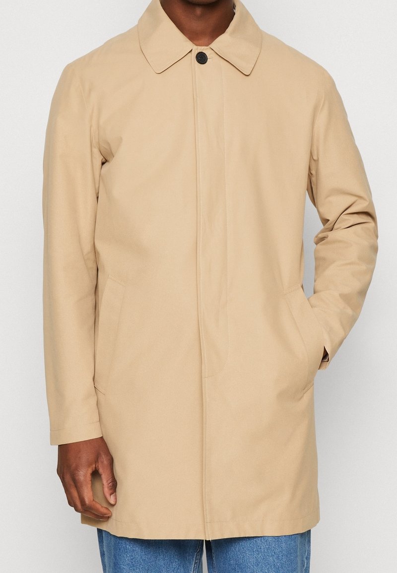 Uomo che indossa un cappotto lungo beige con chiusura a bottoni nascosta e tasche laterali, abbinato a jeans blu, in piedi contro uno sfondo neutro.