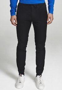 Czarne spodnie dresowe z miękkiego materiału, z paskiem ściągającym w talii, dopasowanym krojem zwężanym ku dołowi, w zestawieniu z białymi trampkami.