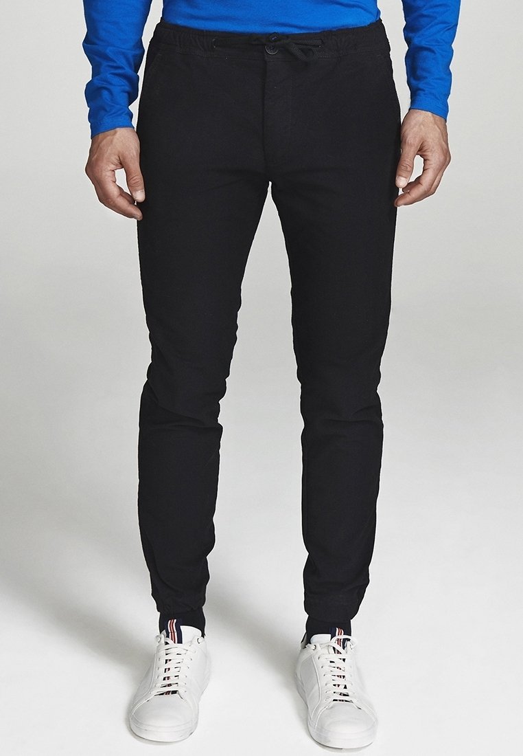 Czarne spodnie dresowe z miękkiego materiału, z paskiem ściągającym w talii, dopasowanym krojem zwężanym ku dołowi, w zestawieniu z białymi trampkami.