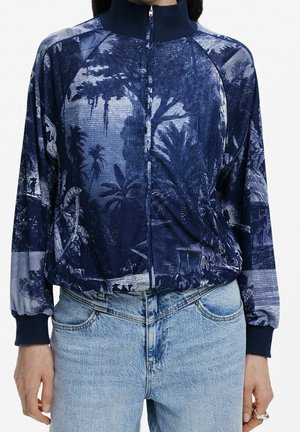 Vrouw draagt een marineblauwe jas met rits met een gedetailleerde blauwe natuur- en bomenlandschapsprint, gecombineerd met lichtblauwe high-waist jeans.