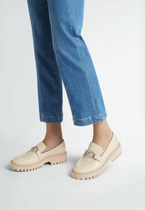 Mocassins beiges avec un dessus brillant, une sangle et une boucle détaillées, combinés avec une semelle épaisse et striée. Associés à un pantalon en denim bleu retroussé.