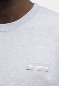 Gros plan d'une personne portant un t-shirt gris clair à col rond avec un petit patch rectangulaire du logo Columbia sur la poitrine.