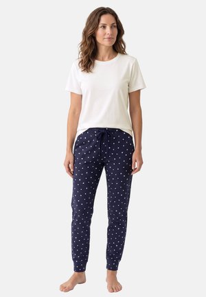 Kvinde står barfodet iført en hvid t-shirt med korte ærmer og marineblå pyjamasbukser med hvide hjerter og snøre i taljen.