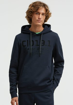 Junger Mann mit lockigem Haar, der einen dunkelblauen Hoodie mit Fronttasche und schwarzem Aufdruck trägt, vor einem schlichten hellen Hintergrund stehend.