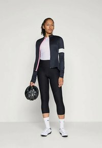 Tengerészkék kerékpáros dzseki rózsaszín akcentussal, világos rózsaszín felső, fekete leggings, és fehér zoknik. A modell egy fekete sisakot tart, semleges környezetben áll.