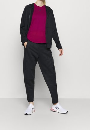 Černá mikina na zip, magentové tričko, černé kalhoty se vzorem a bílé sportovní boty s červenými akcenty. Součástí jsou kapsy a logo.