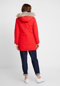 Rode geïsoleerde parka met een capuchon gevoerd met namaakbont, lange mouwen en een lengte tot halverwege de dij, gecombineerd met opgerolde donkere jeans en witte sneakers.