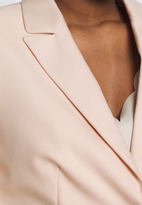 Blazer rose clair, tissu lisse, coupe ajustée, revers crantés nets, décolleté en V profond visible, détails de couture minimaux.