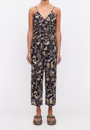 Musta haalari, jossa on keltainen ja violetti paisley-kuosi, V-mallinen kaula-aukko, gathered-rintaosa ja leveät, lyhyet lahkeet. Mallilla on jaloissaan litteät sandaalit.