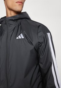 Sort letvægtsjakke med lynlås foran, hætte og hvide diagonale striber på ærmerne. Har et Adidas-logo på brystet.