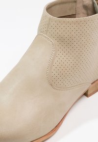 Beiger Leder-Stiefel bis zum Knöchel mit perforierten Details, gebogenem Schaft und niedrigem Holzabsatz, der eine glatte Textur und ein minimalistisches Design zeigt.