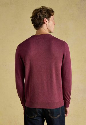 Man met kort krullend haar, gekleed in een bordeauxrode trui met lange mouwen en donkere spijkerbroek, staand met de rug naar een effen beige achtergrond.
