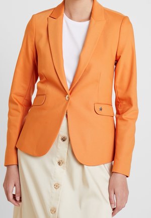 Femme portant un blazer orange par-dessus une chemise blanche et une jupe beige avec des boutons sur le devant, sur un fond uni.