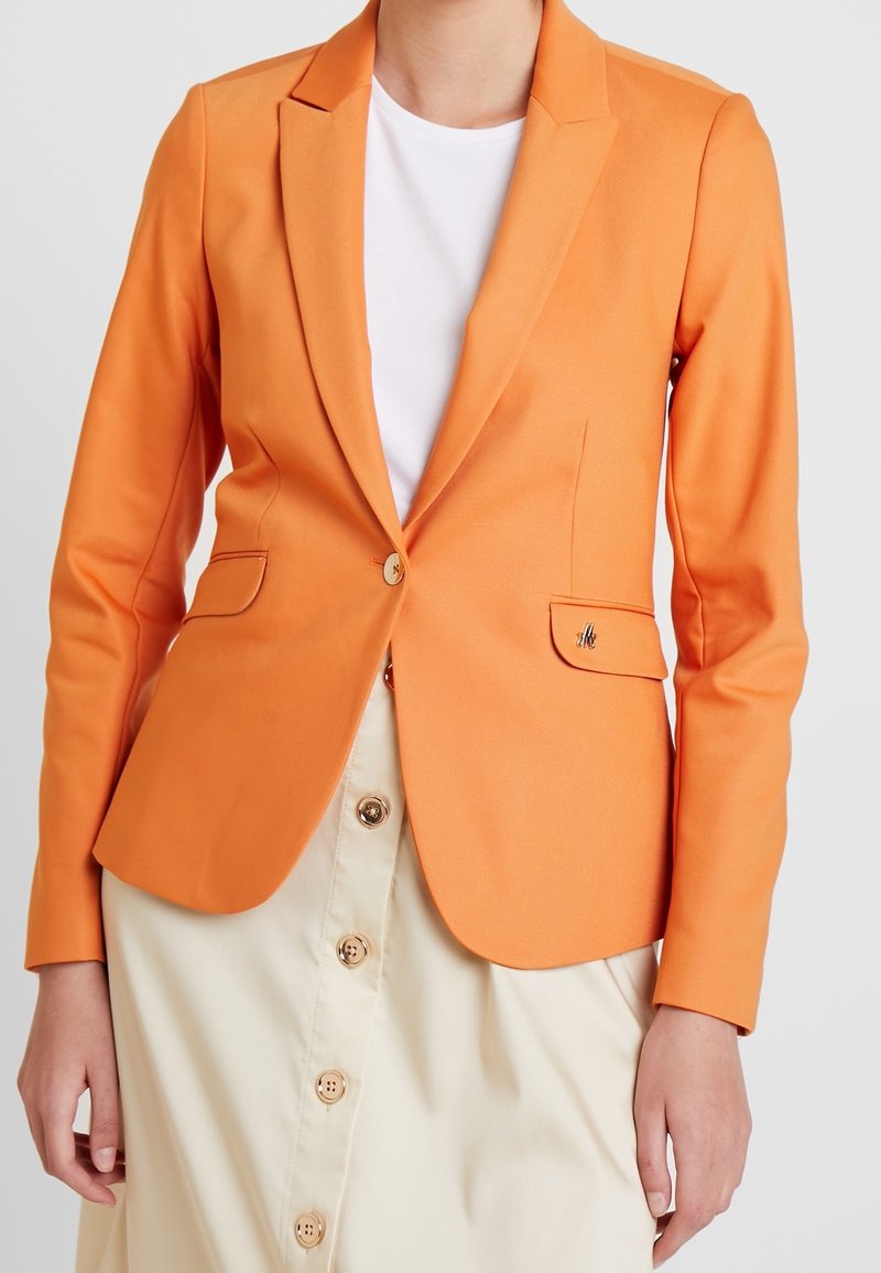 Femme portant un blazer orange par-dessus une chemise blanche et une jupe beige avec des boutons sur le devant, sur un fond uni.