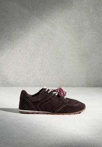 Baskets en daim marron foncé avec œillets blancs, lacets blancs et roses noués en nœud, et semelle en caoutchouc marron clair sur une surface grise.