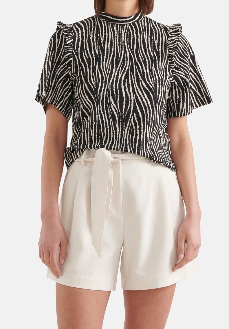 Femme portant un top à manches courtes rayé zèbre noir et blanc avec des détails à volants, associé à un short blanc taille haute avec une ceinture nouée à l'avant.