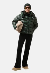 Grøn pufferjakke med en glat finish, kombineret med sorte bukser og beige sneakers. Tilbehøret omfatter en strikket beanie og store solbriller.