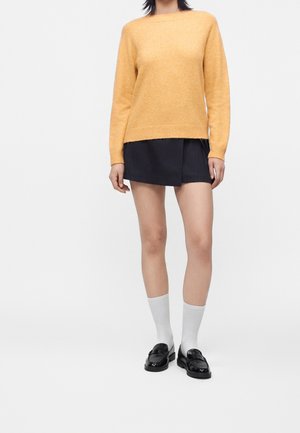 Femme portant un pull jaune, une mini-jupe noire, des chaussettes blanches et des mocassins noirs, debout sur un fond clair.