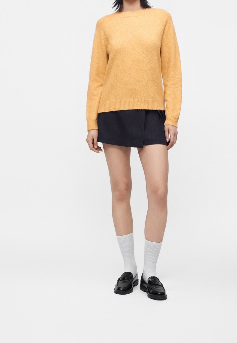 Femme portant un pull jaune, une mini-jupe noire, des chaussettes blanches et des mocassins noirs, debout sur un fond clair.