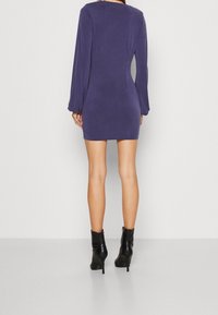 Fialové mini šaty s dlouhým rukávem, upnutým střihem a jemnou texturou, s kulatým výstřihem a mírně rozšířenými manžetami. Černé kotníkové boty.